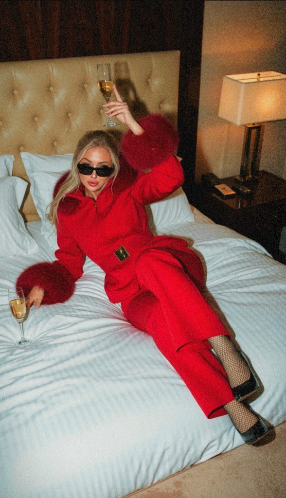 Amber Della Vita in a red ski suit on a hotel bed with wine